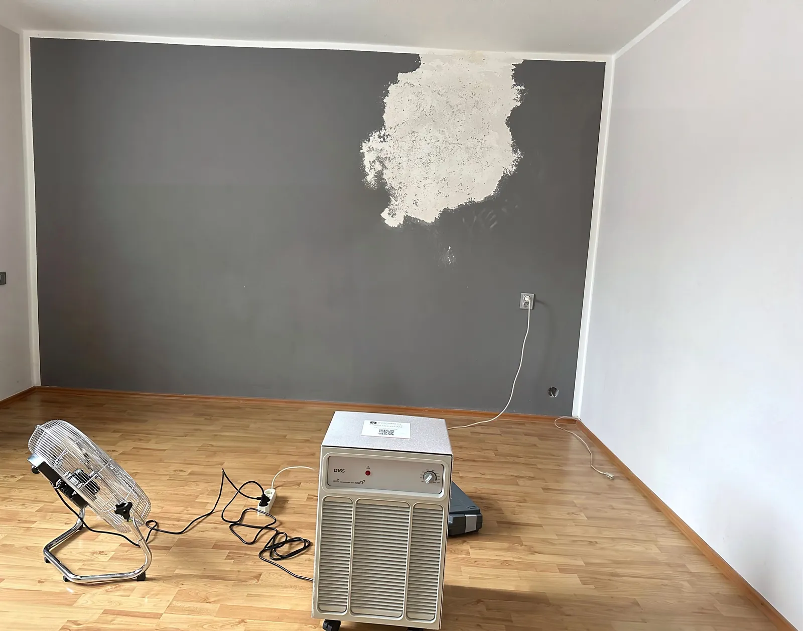 Kondenzační vysoušeč a ventilátor v ložnici u zdi s oškrábanou omítkou.